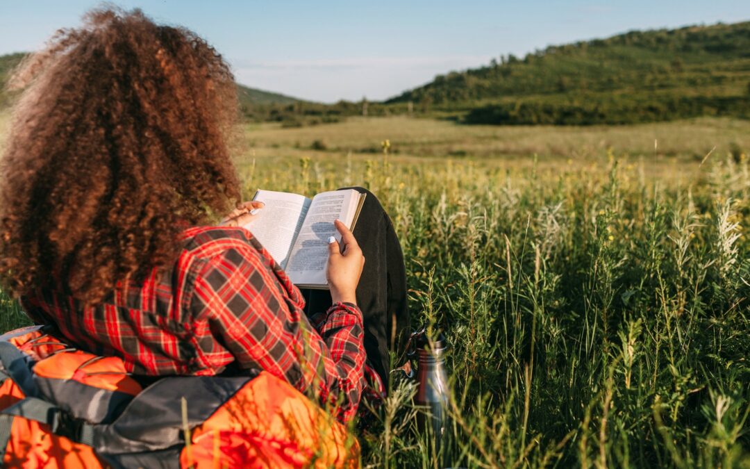 A woman with her back to the camera, sitting in a field reading a book.