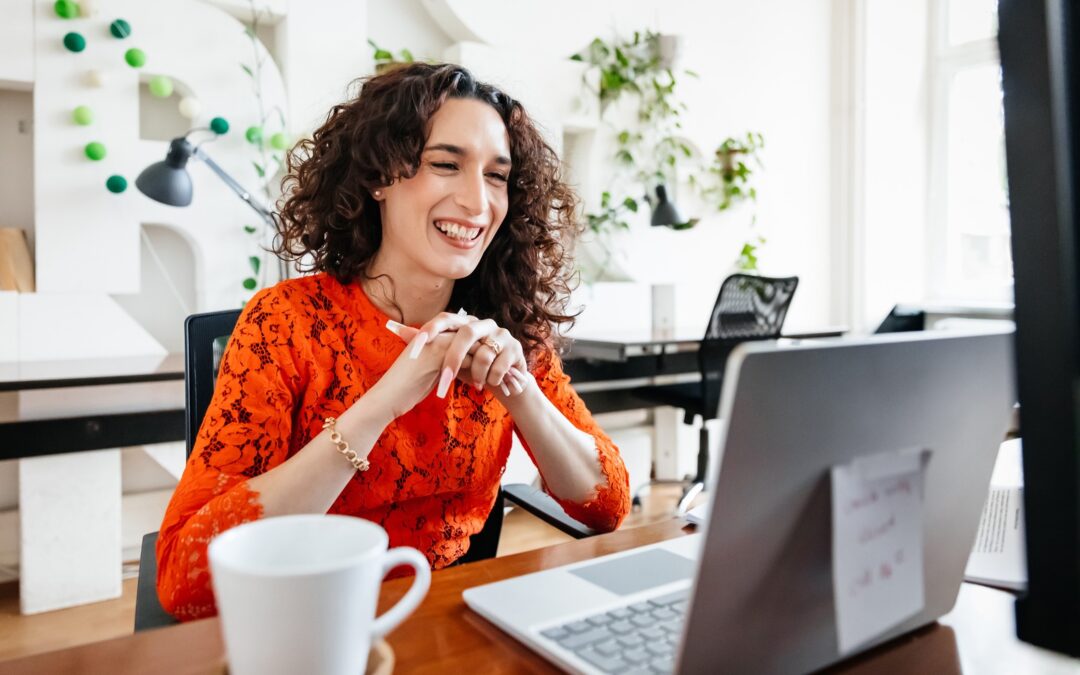 A middle-aged woman with curly dark hair and wearing an orange shirt sits at a table an smiles at her open laptop