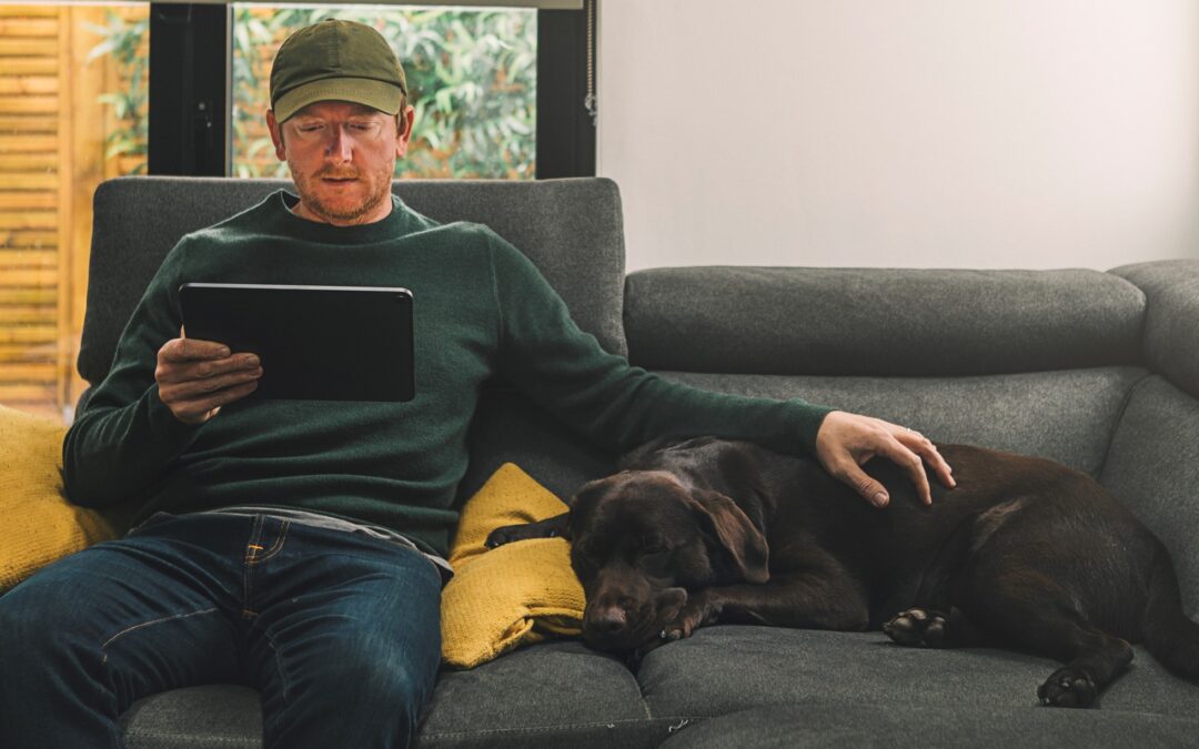 A man sits on his couch looking at a tablet while petting his dog